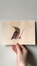 'Owl' print held in a hand: an owl in a green knitted hood holding a sword