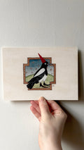 'Magpie' print held in a hand: a magpie in a red pointed hat standing on a green hill in a stepped frame