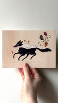 'The Hunt' print held in a hand: a black wolf carrying a white bird in its mouth, beside oak leaves