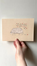 'Grazing Creacher' print held in a hand: a woolly white sheep-like creacher grazing under red flowers