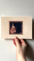 'Strawberry Lunch' print held in a hand: a pink horned creacher eating a strawberry, on dark blue in a border
