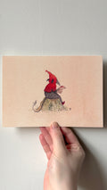 'Quarlic Tom Reads a Letter' print held in a hand: a creature in a red pointed hood reading on a mossy rock