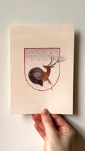 'Sneer, the Snail-Deer' print held in a hand: a deer with a snail shell body inside a shield shape