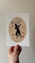 'The Shade' print held in a hand: a black antlered creature playing a violin in an oval of stars