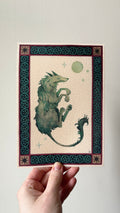 "Cù-Sìth" print held in a hand: a green shaggy dog leaping under a pale moon, in a green knotwork border