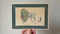 "Each-Uisge" print held in a hand: a pale horse-like water spirit with a weedy mane, in a green border