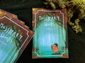 "Mossberry Woods" art book: the teal cover with a small creature in a forest, beside another copy on moss