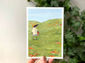 "Summer Birdface" print held in a hand: Birdface in a straw hat carrying a basket through a green meadow