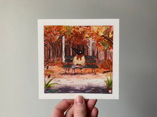 "Autumn" print held in a hand: Birdface sitting on a park bench among orange autumn trees