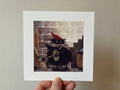 "Soren Spider Stealer I" print held in a hand: Soren, a black furry creature in a red pointed hat, in a cellar