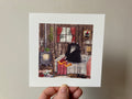 "Gremble's Socks" print held in a hand: a black furry creature sitting on a bed in a wooden cabin