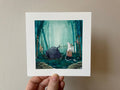 "The Cauldron" print held in a hand: a white creature with a basket beside a fallen cauldron in a teal forest