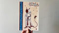 "The Beekeeper" print held in a hand: Silence Plimsole, a robed figure in a beekeeper's veil, by a blue border