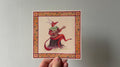 "The Lutenist" print held in a hand: a red fox-like creature with antlers playing a lute, in an orange border