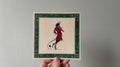 "The Fiddler" print held in a hand: a black long-eared creature in a red dress playing a fiddle, green border