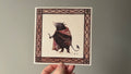 "The Forager" print held in a hand: a mole-like creature in a pink cloak inside a brown patterned border